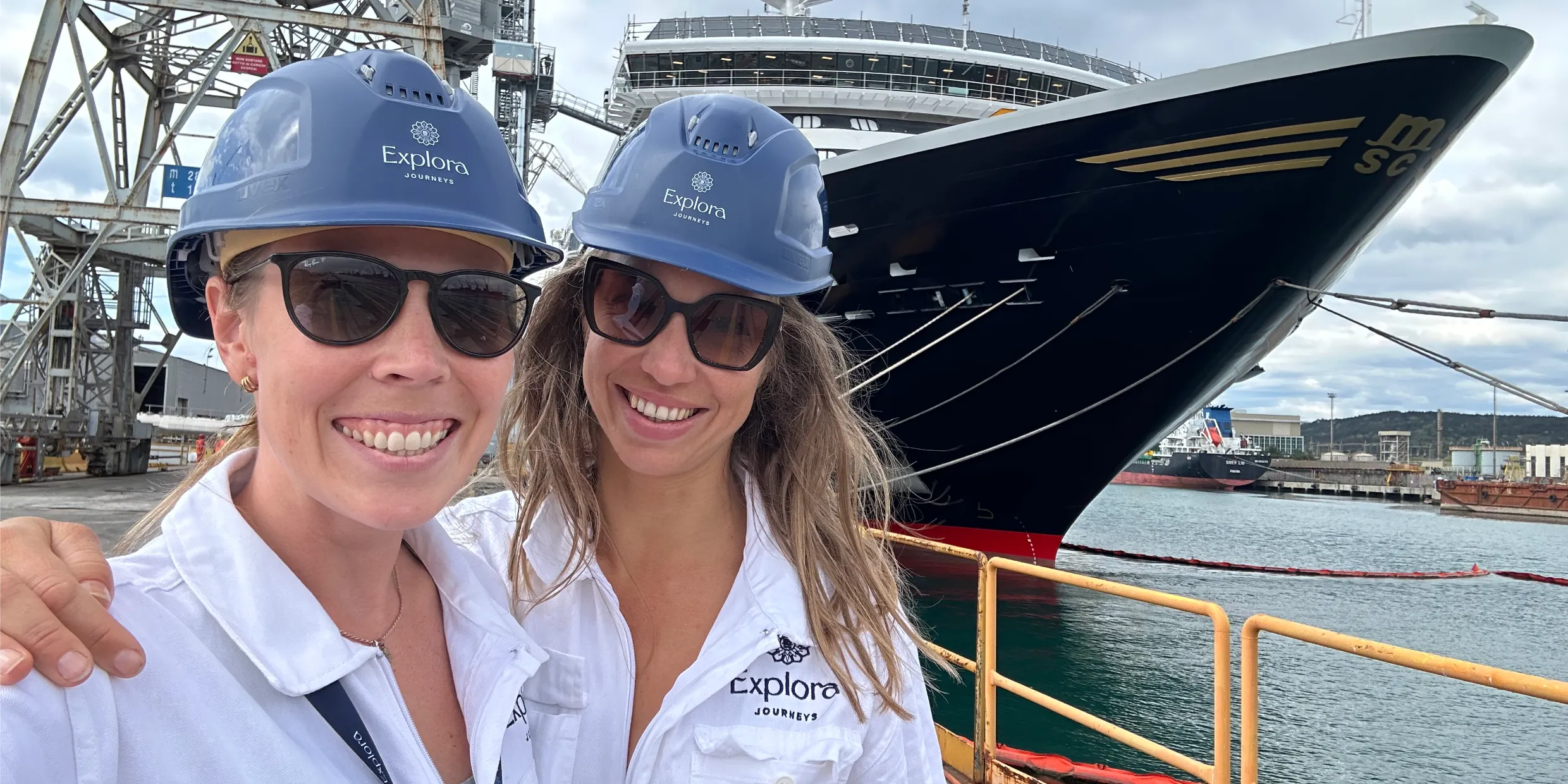 Two colleagues in hard hats and protective gear on a shipyard quayside beside a moored vessel.