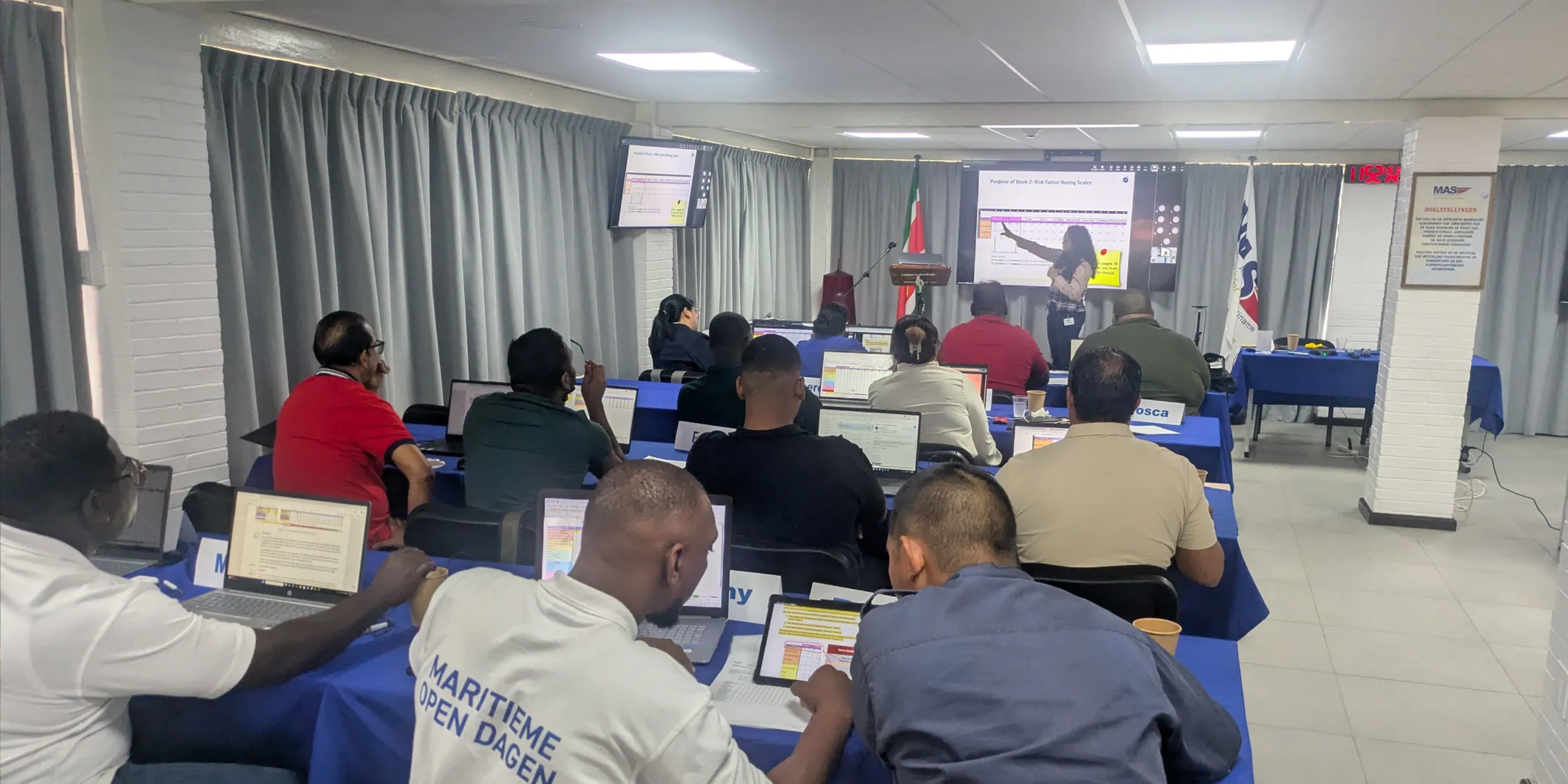 A woman presents risk-assessment material at a screen to a room of port professionals working at laptops.